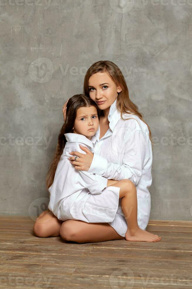 intérieur portrait de une magnifique mère avec sa charmant peu fille posant contre une gris mur. photo