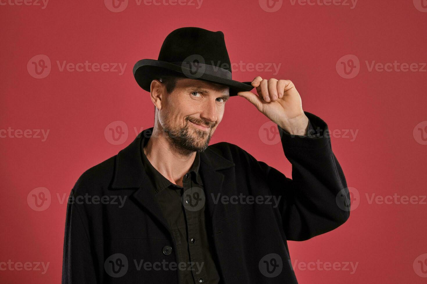 âge moyen homme avec barbe et moustache, porte noir chapeau et veste posant contre une rouge Contexte. sincère émotions concept. photo