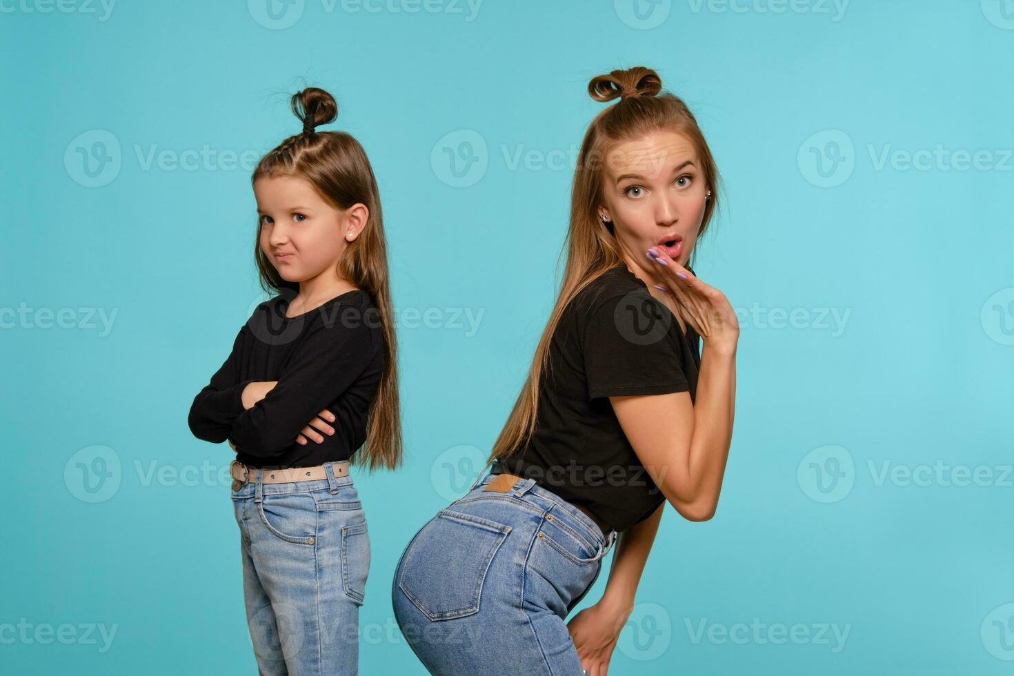 maman et fille avec une marrant coiffures, habillé dans noir chemises et bleu denim jeans sont posant contre une bleu studio Contexte. fermer tir. photo