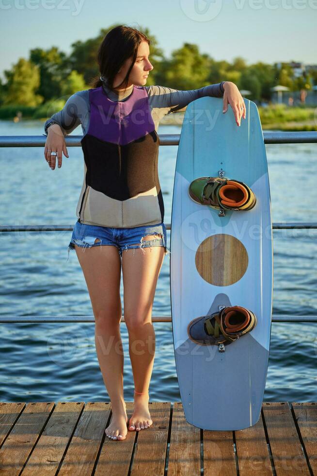 fille dans une gris col roulé, spécial des sports gilet et bleu denim short est à la recherche une façon tandis que posant avec sa wakeboard sur une jetée. plein longueur tir. photo