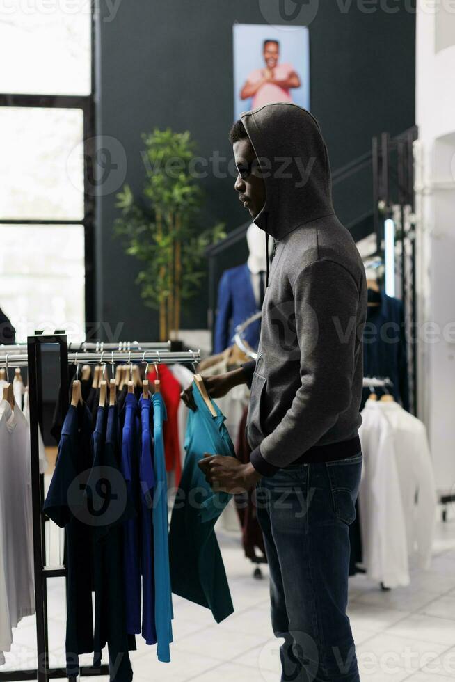 africain américain homme à la recherche de côté tandis que voler Vêtements magasin, vérification racks avec branché vêtements. voleur en essayant à voler à la mode marchandise, portant des lunettes de soleil et capuche ne pas à être reconnu photo