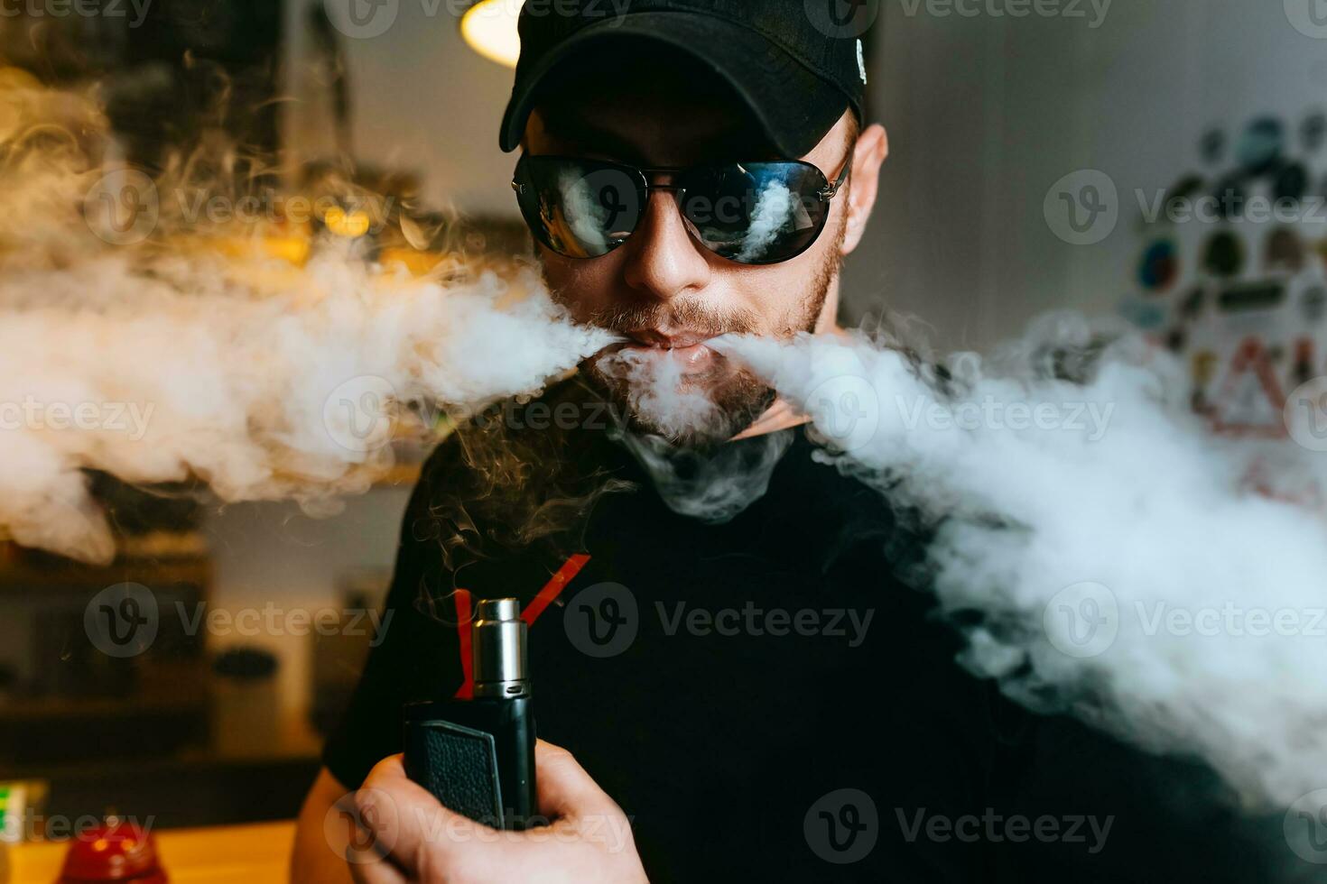 brutal barbu homme dans des lunettes de soleil fumée un électronique cigarette et communiqués des nuages de vapeur à le vape magasin. photo