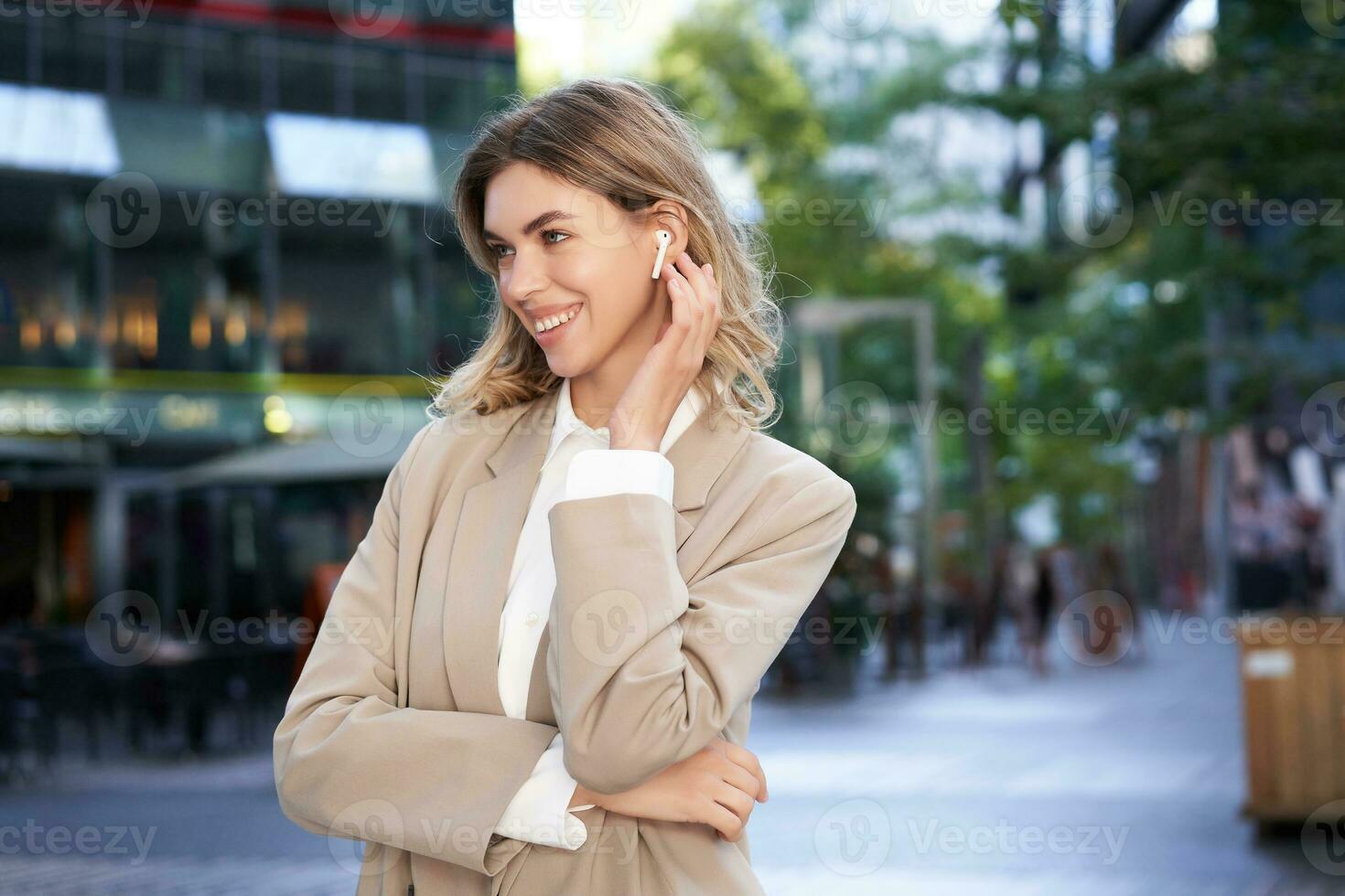 proche en haut portrait de blond femme d'affaires, sur de soi entreprise femme dans écouteurs et beige costume, posant en plein air sur rue de ville centre photo