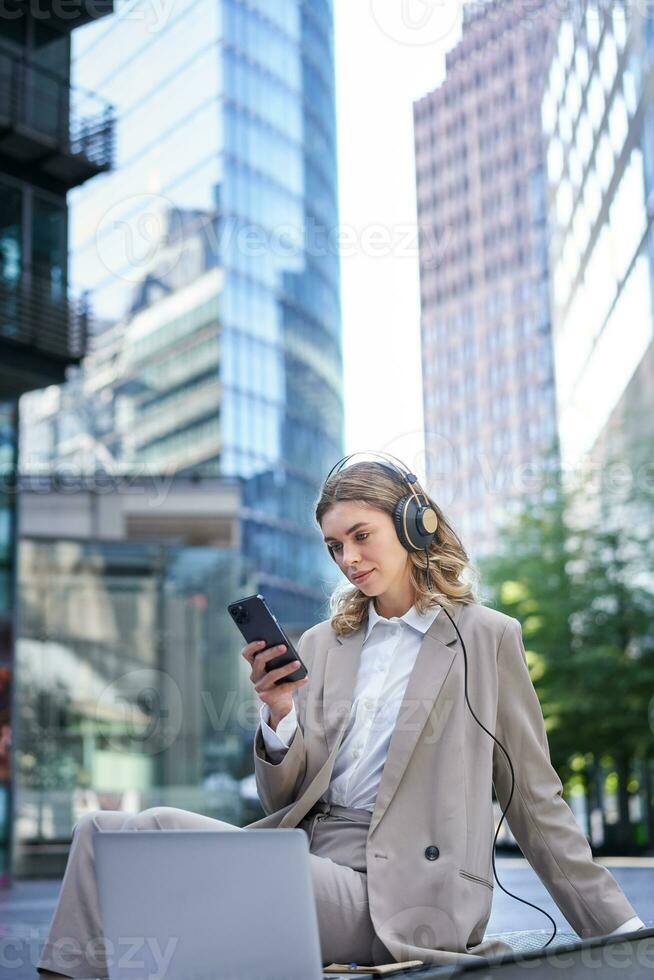 entreprise femme dans costume, séance dans ville, écoute à la musique avec ordinateur portable, défilement nouvelles sur sa mobile téléphone, relaxant sur le déjeuner Pause photo