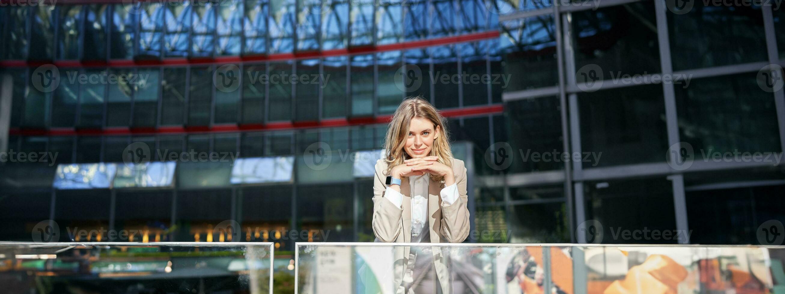 magnifique Jeune femme dans beige costume, permanent près Bureau bâtiments dans ville centre, souriant et à la recherche rêveur photo