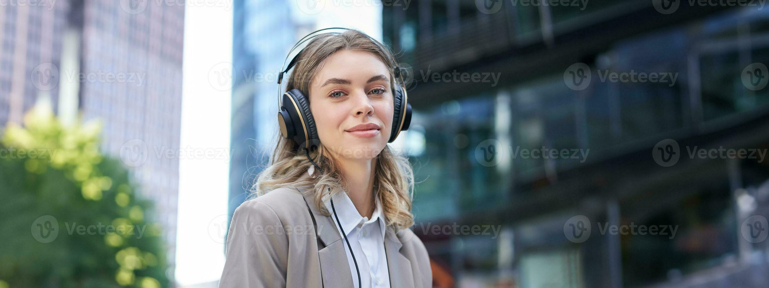 portrait de souriant entreprise femme, fille dans costume écoute la musique dans écouteurs, séance dans ville centre photo