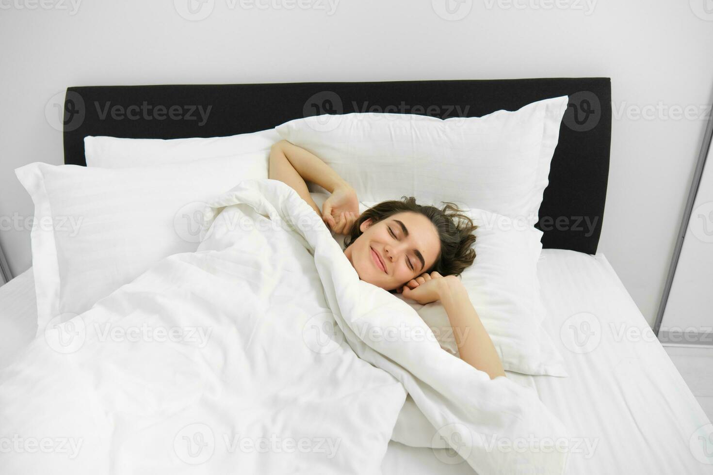 portrait de souriant, heureux Jeune femme élongation sa bras, veille en haut après bien nuit dormir, mensonge sur doux blanc oreiller dans lit avec confortable lin feuilles photo