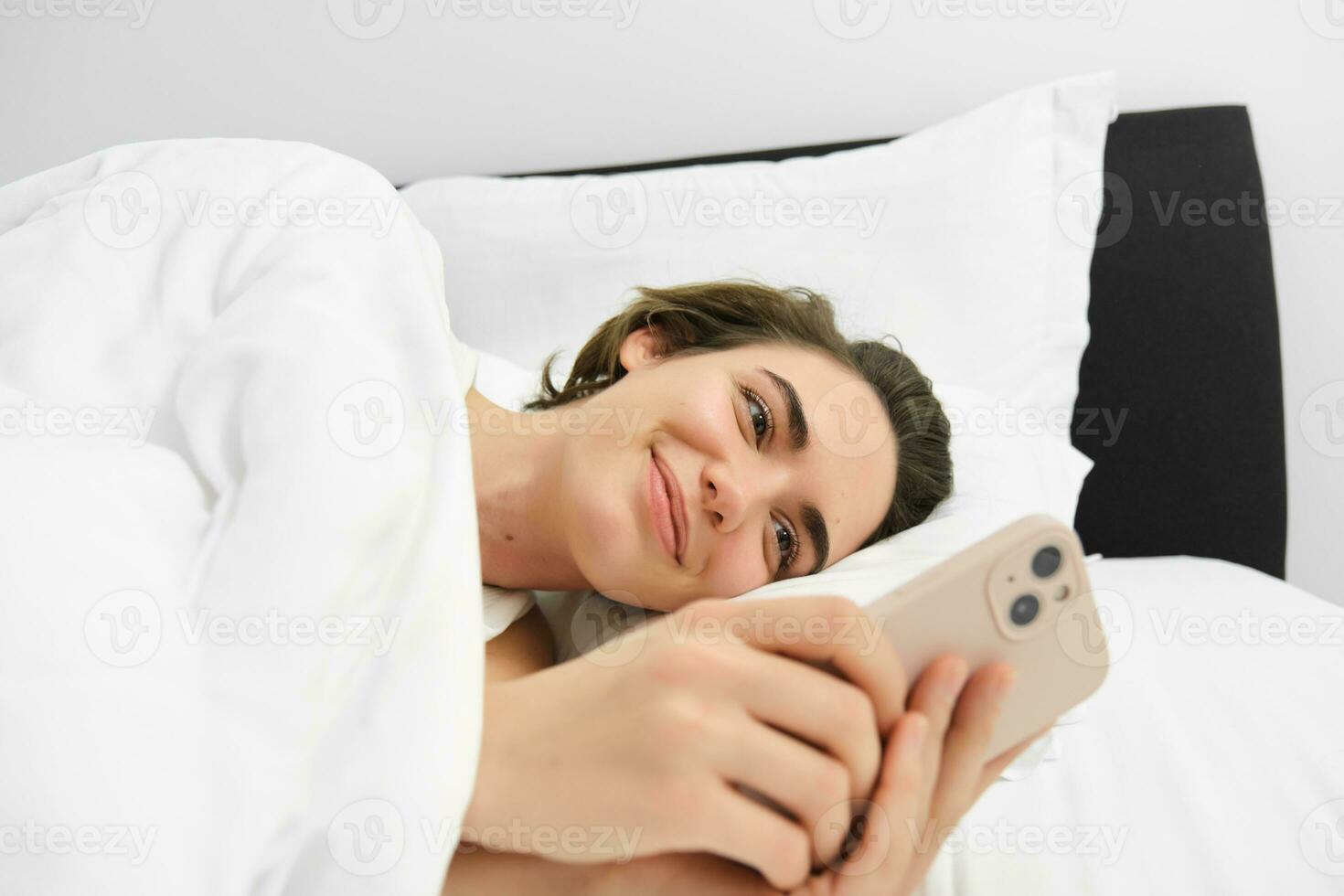proche en haut portrait de charmant Jeune femme dans lit, mensonge et Aller à dormir, vérification sa téléphone messages, en utilisant téléphone intelligent et souriant photo