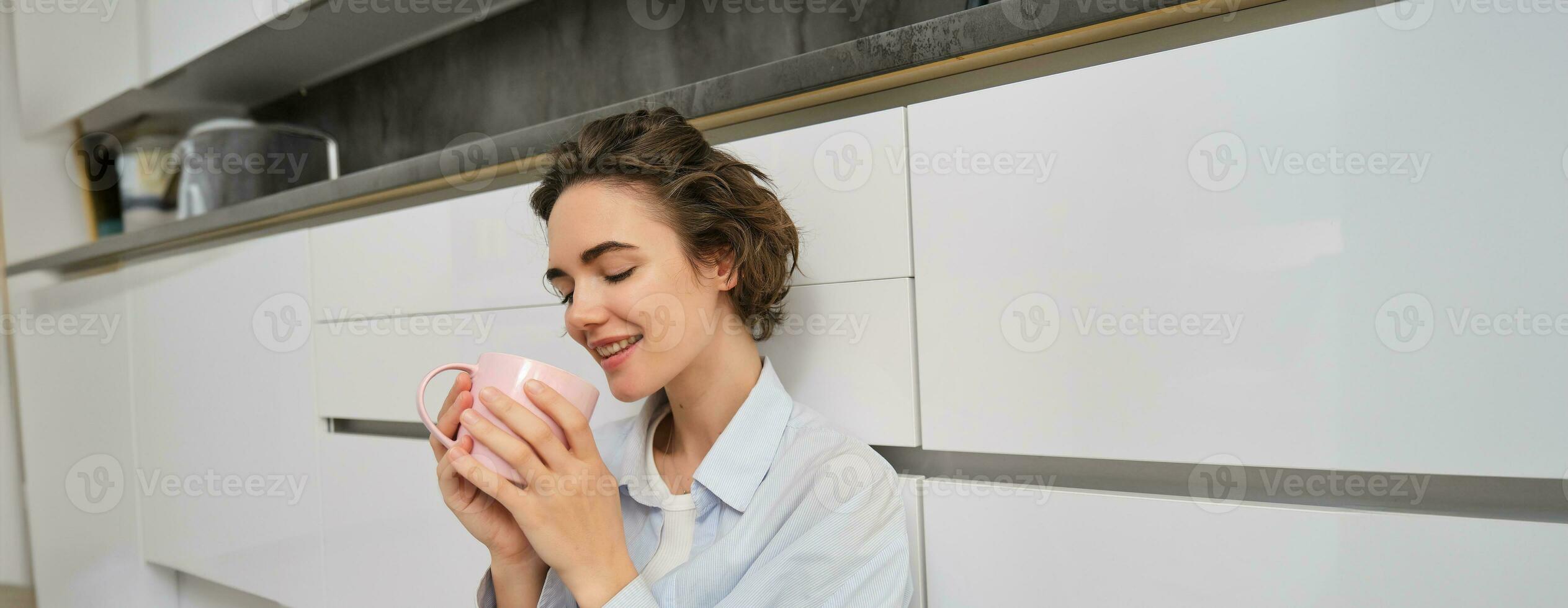 beauté et femmes concept. détendu Jeune femme jouit sa tasse de thé. fille est assis sur sol dans cuisine avec rose tasse, les boissons café et sourit de délice photo