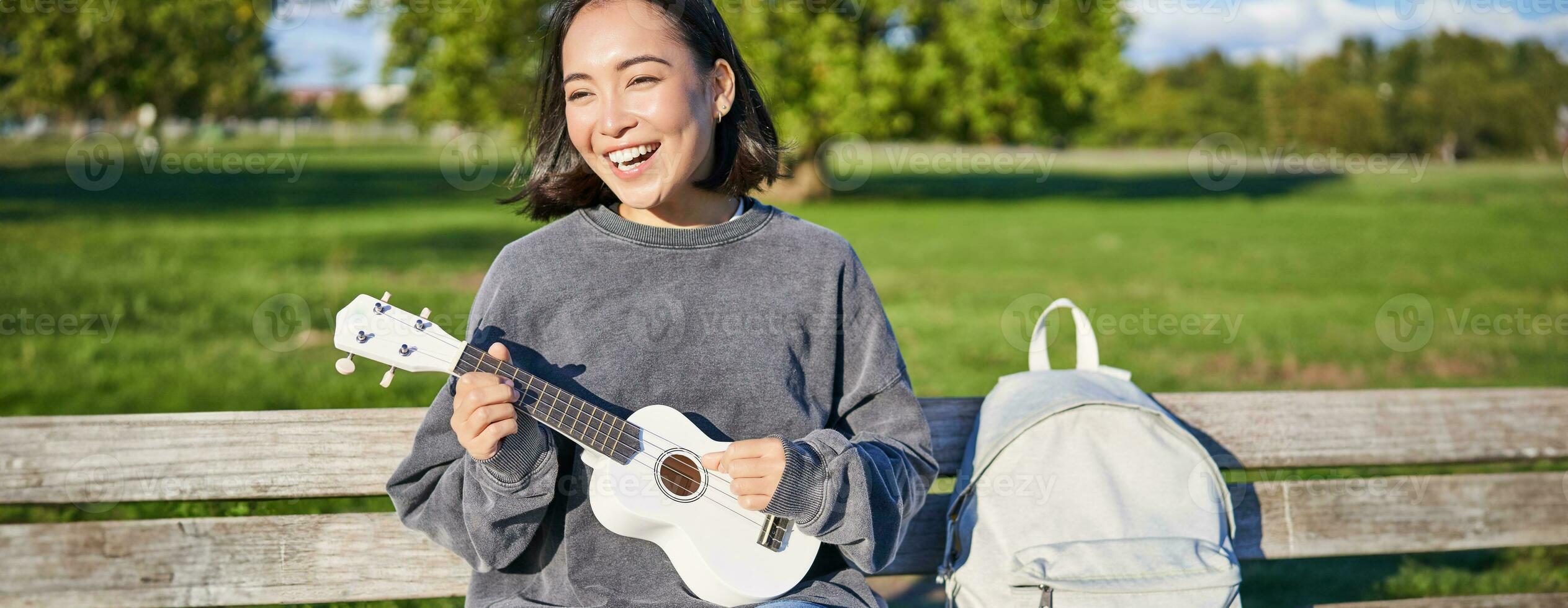 content mignonne fille est assis seul sur banc dans parc, pièces ukulélé guitare et jouit ensoleillé journée en plein air photo