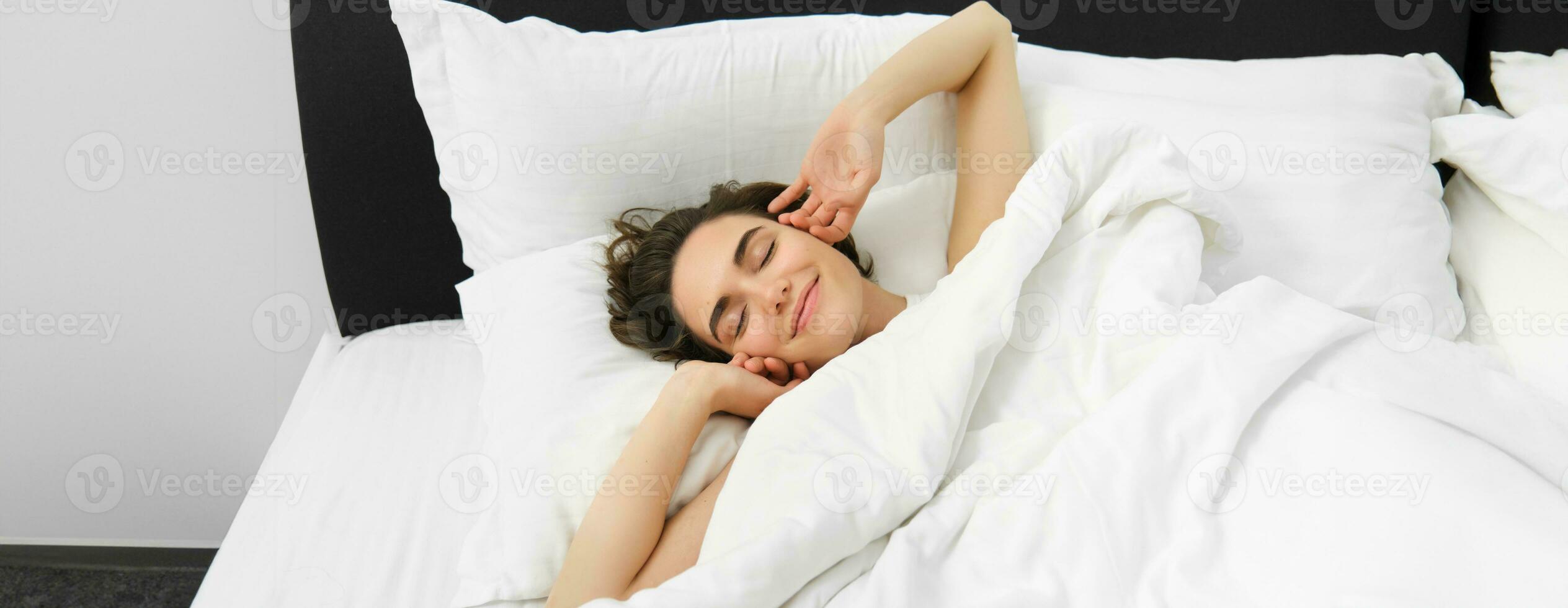 portrait de souriant, heureux Jeune femme élongation sa bras, veille en haut après bien nuit dormir, mensonge sur doux blanc oreiller dans lit avec confortable lin feuilles photo