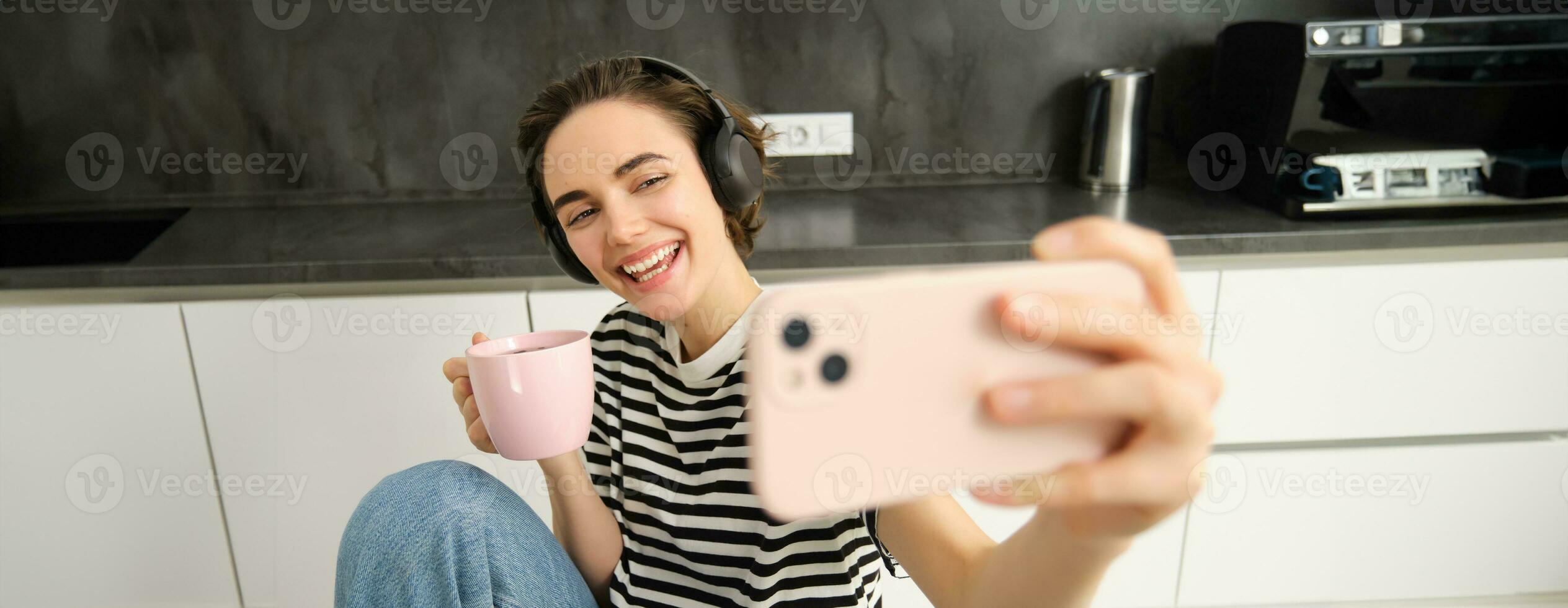 proche en haut portrait de femme avec téléphone intelligent, prise selfie tandis que en buvant thé et écoute la musique dans écouteurs, fabrication Photos pour sa social médias app dans le cuisine