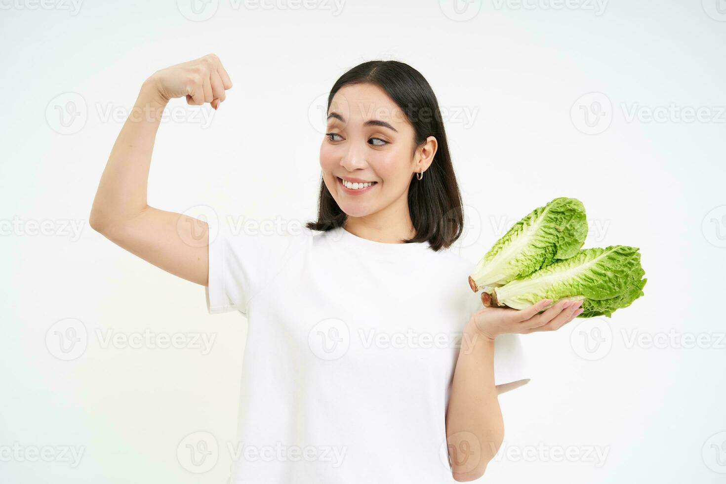 fort asiatique femme spectacles Frais légumes, salade et sa muscles, fléchissement biceps avec souriant affronter, blanc Contexte photo