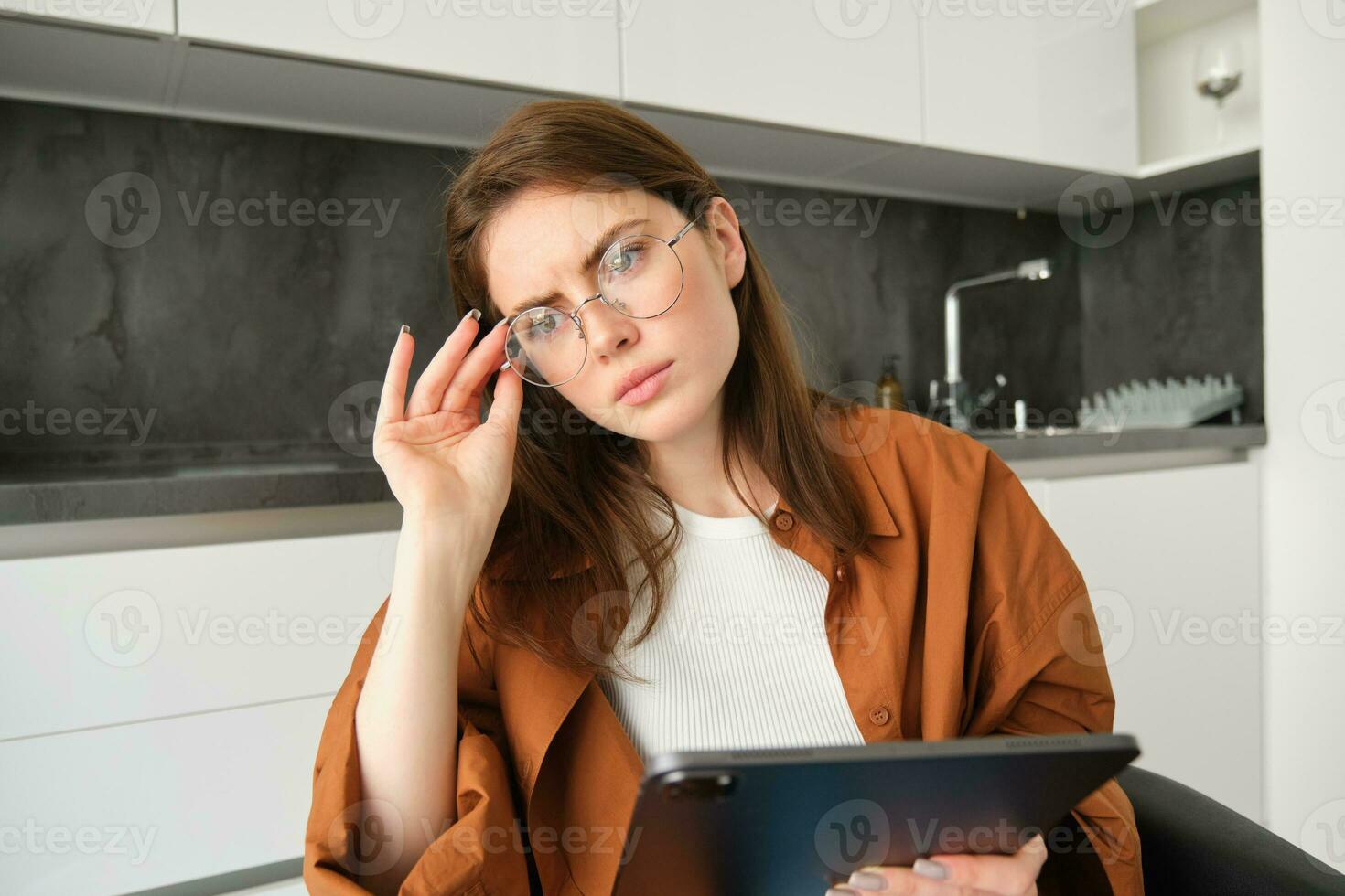 portrait de sérieux Jeune femme dans lunettes, en portant tablette, séance dans cuisine, à la recherche une façon dans Profond pensées, pensée, a concerné visage photo