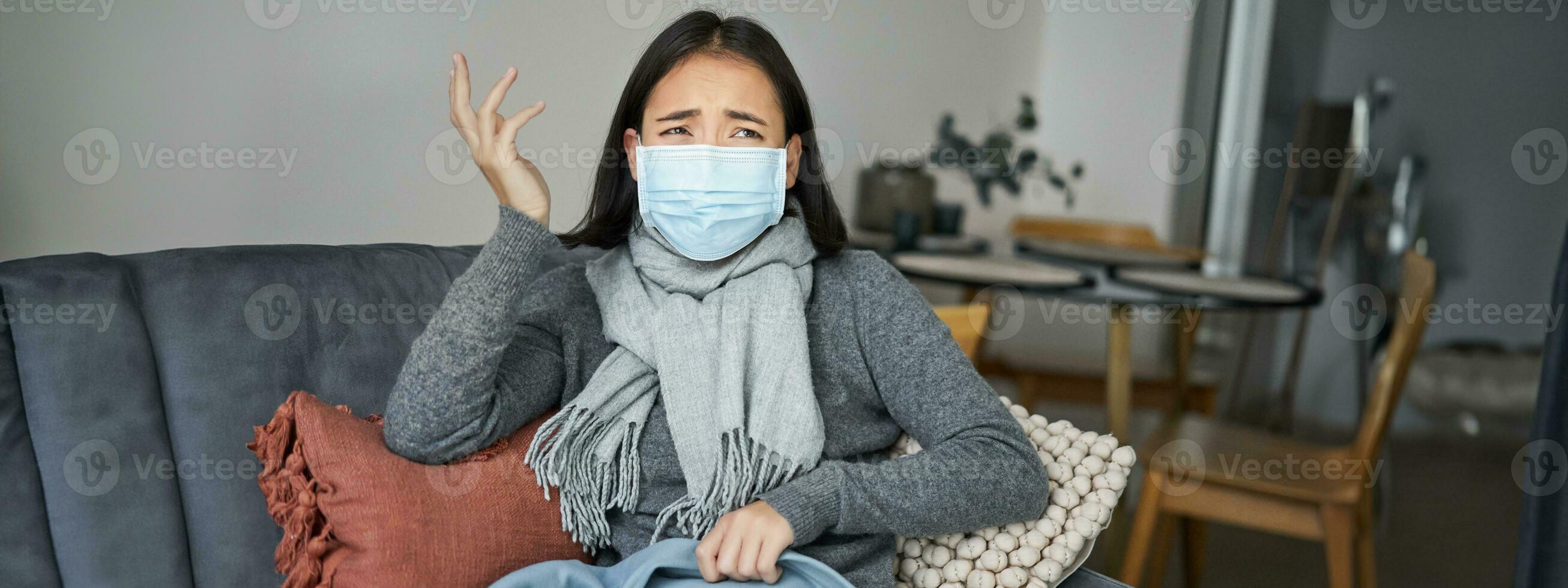 covid-19 et santé concept. Jeune asiatique femme dans médical visage masque, se sent malade et indisposé, contagieux grippe, protéger autres de grippe, séance dans vivant pièce couvert avec couverture photo