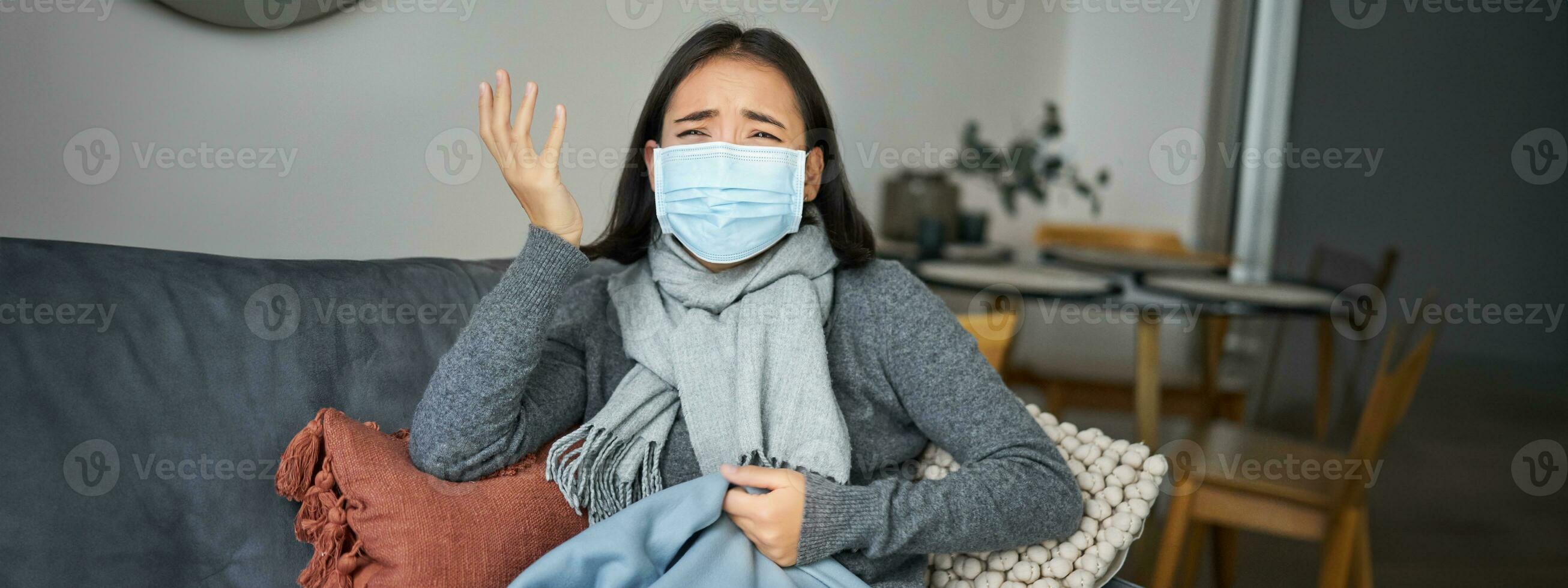 covid-19 et santé concept. Jeune asiatique femme dans médical visage masque, se sent malade et indisposé, contagieux grippe, protéger autres de grippe, séance dans vivant pièce couvert avec couverture photo