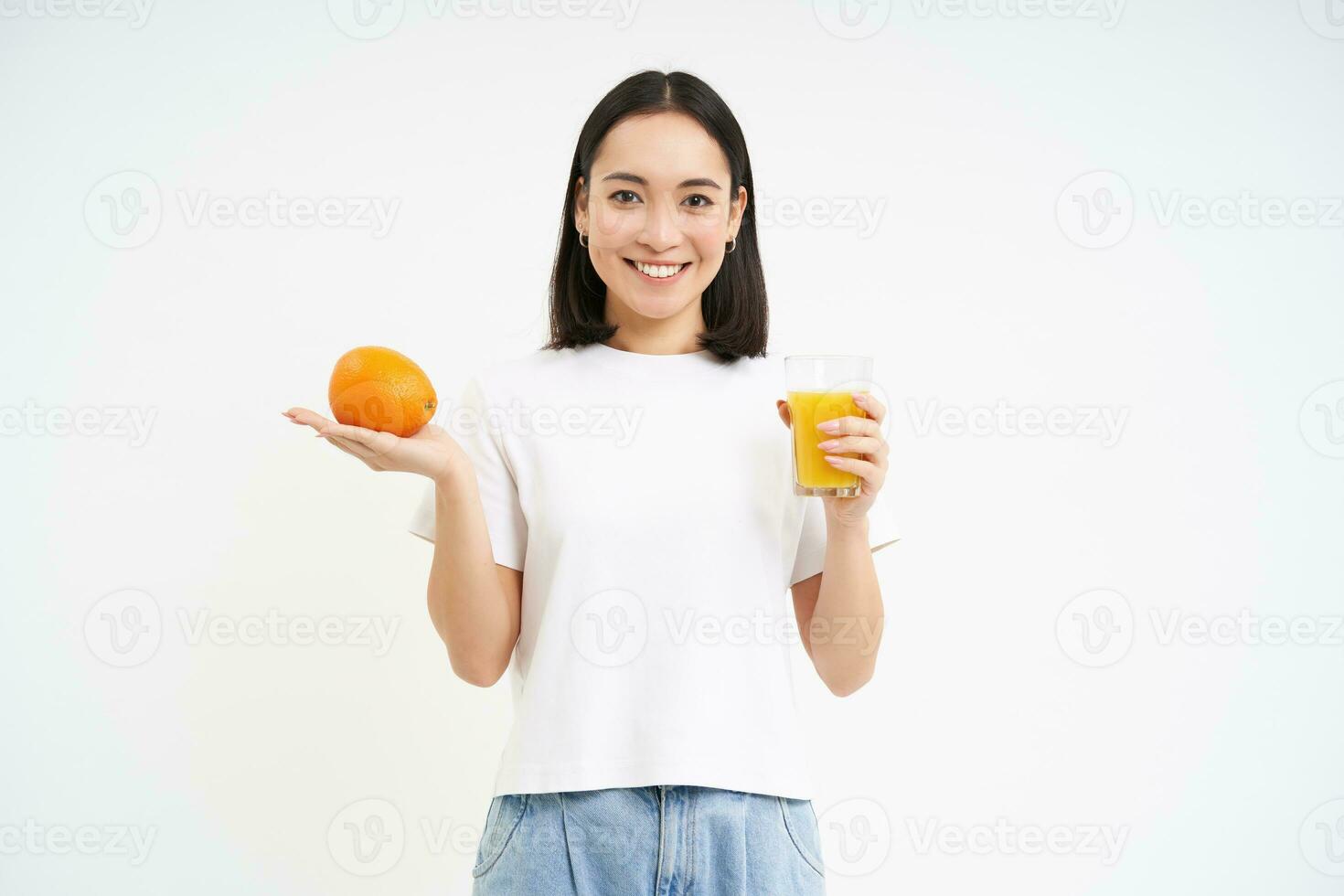 bien-être et en bonne santé mode de vie. souriant asiatique femme montrant Orange fruit et jus, serre Frais boire, permanent plus de blanc Contexte photo