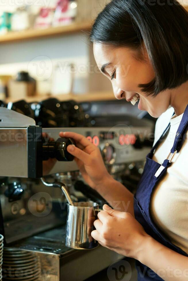 content asiatique femme, barista en utilisant café machine à faire commande, fumant Lait pour cappuccino et latté, en riant et souriant tandis que travail dans café photo