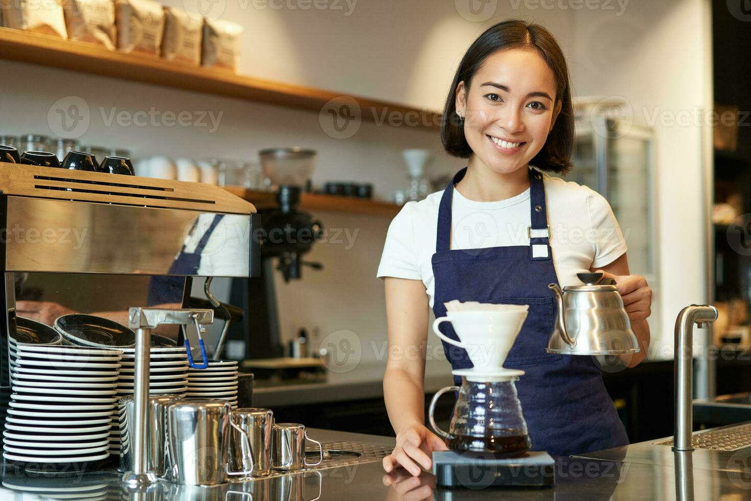 souriant fille barman, asiatique barman verser l'eau de bouilloire, brassage filtre café dans café derrière comptoir, portant bleu tablier photo