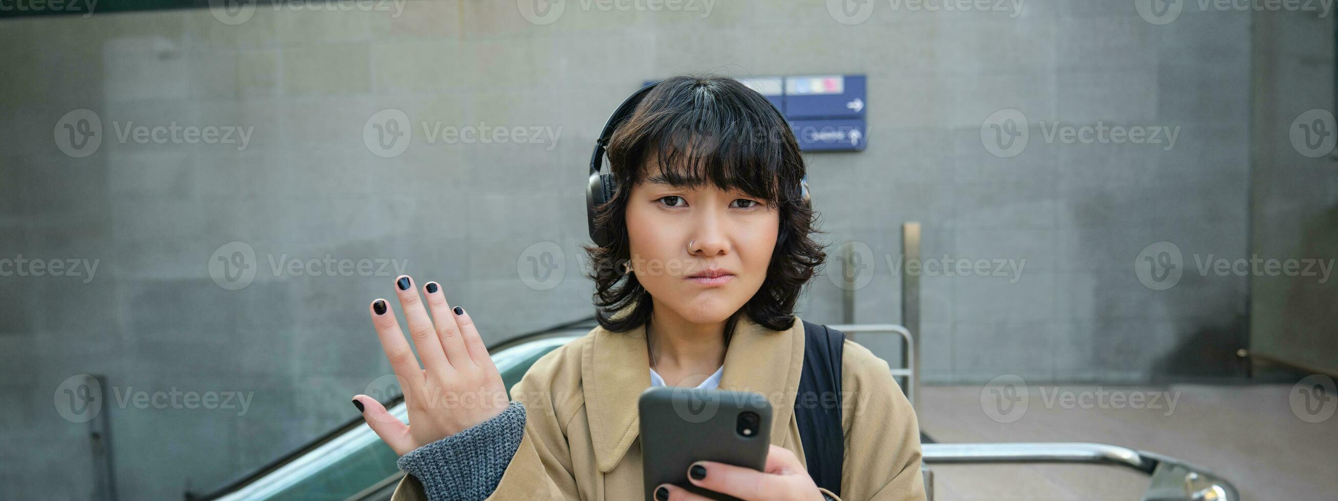 portrait de asiatique fille dans écouteurs, regards compliqué à téléphone intelligent filtrer, perplexe par texte message ou notification, des stands sur rue et hausse les épaules photo