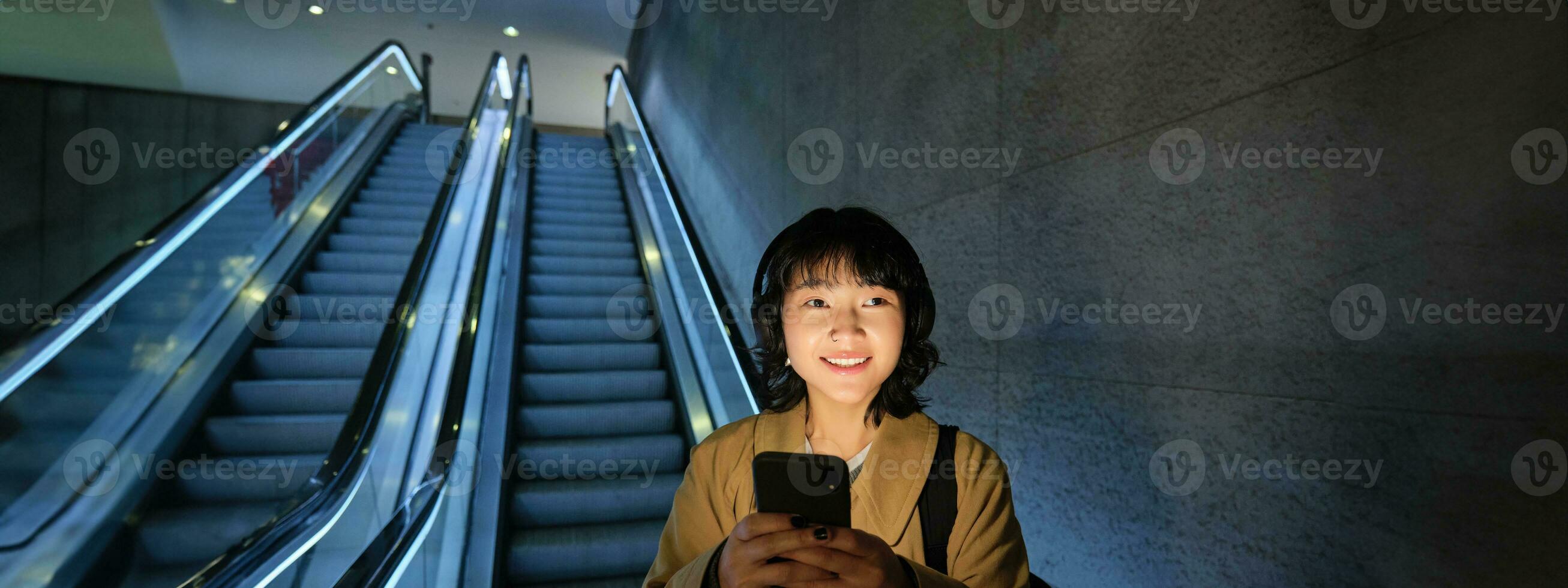 mignonne Jeune femme Aller vers le bas le escalier mécanique à le tube, en utilisant métro métro à commuer à travail ou université, permanent avec téléphone intelligent photo