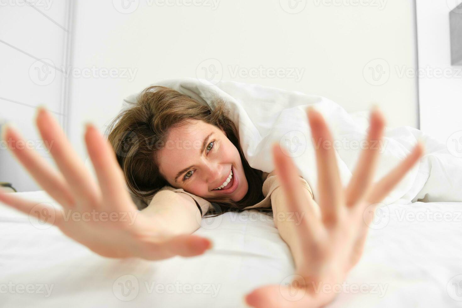 magnifique femme mensonge dans lit, élongation sa bras vers caméra et en riant, veille en haut dans Matin dans content ambiance photo