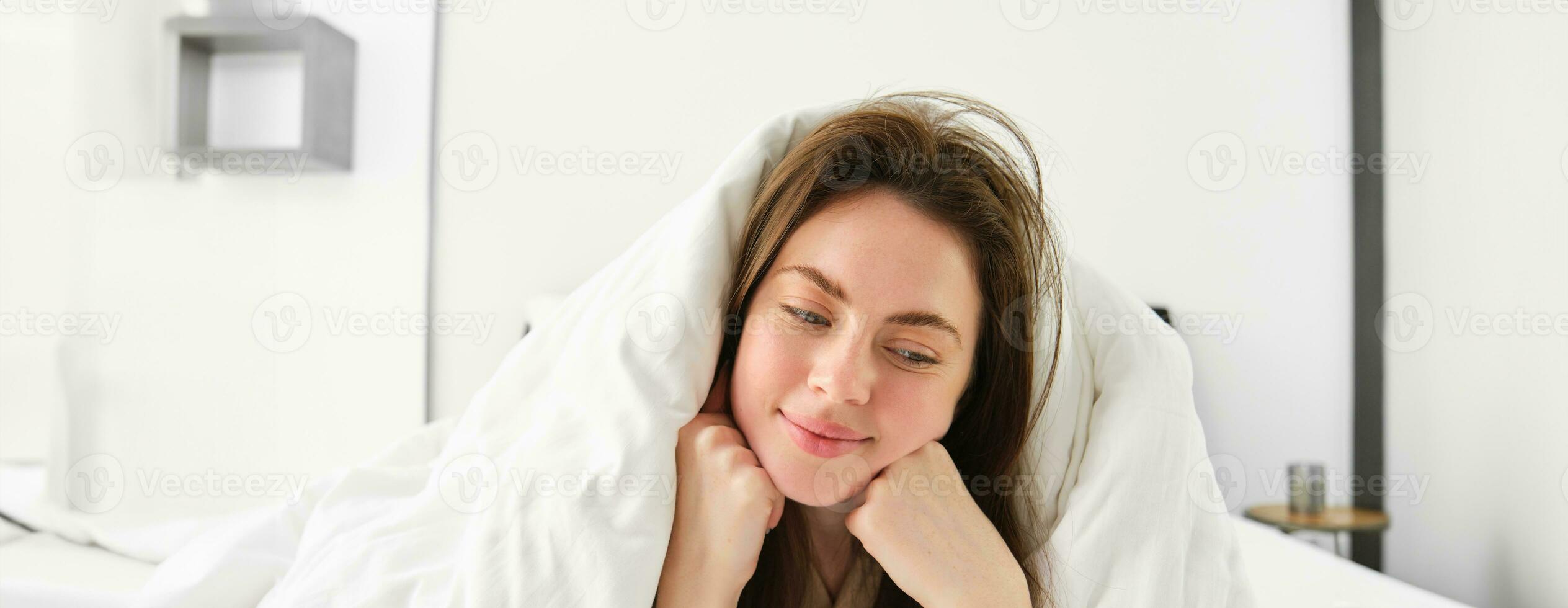 mignonne fille avec désordonné cheveux, mensonge dans lit couvert dans blanc feuilles couette, souriant et en riant coquette, dépenses temps dans sa chambre photo