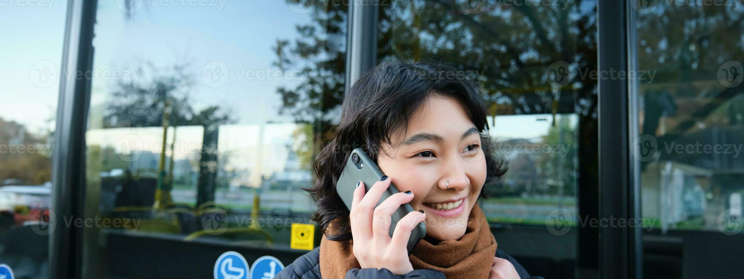 cellulaire La technologie et gens concept. élégant asiatique fille pourparlers sur mobile téléphone, fait du une Téléphone appel, des stands près autobus Arrêtez et a conversation photo