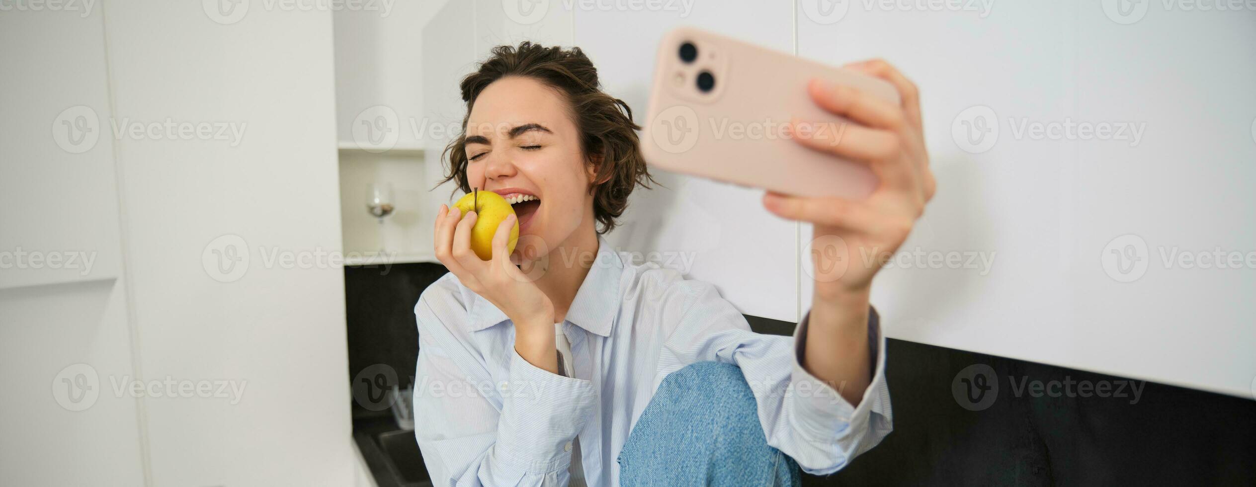 Candide, content Jeune femme mordant un Pomme pour une selfie, prend photo sur téléphone intelligent tandis que en mangeant fruit