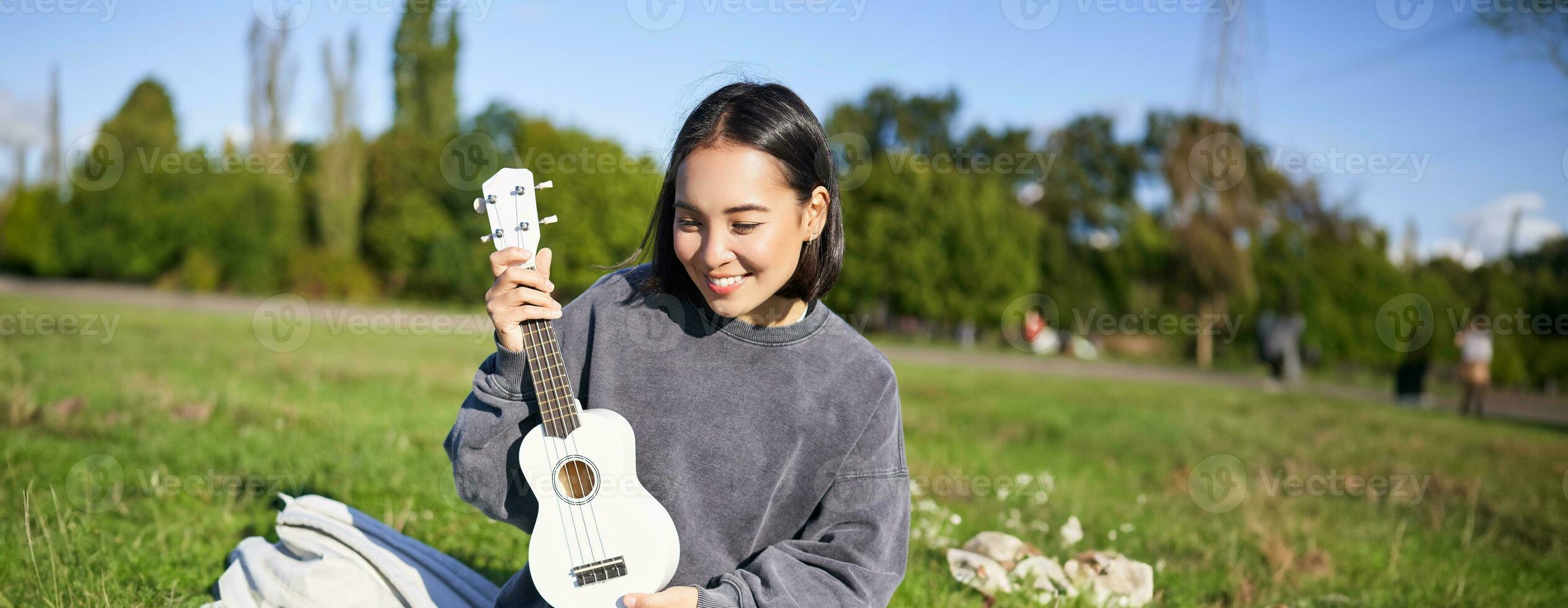 verticale coup de content asiatique fille pièces instrument, spectacles sa ukulélé à portable caméra, vidéo chats à propos musique, enseigne Comment à jouer, est assis dans parc en plein air photo