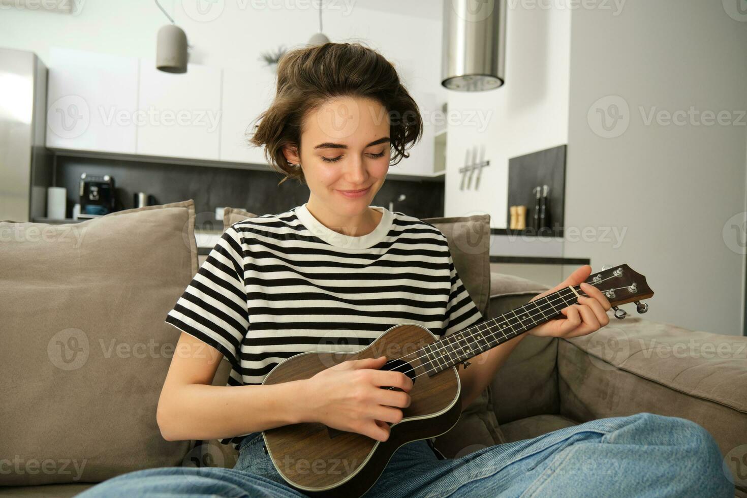 portrait de Jeune moderne femme, étudiant en jouant ukulélé à maison, séance avec petit guitare, en chantant et sentiment content, séance sur canapé photo