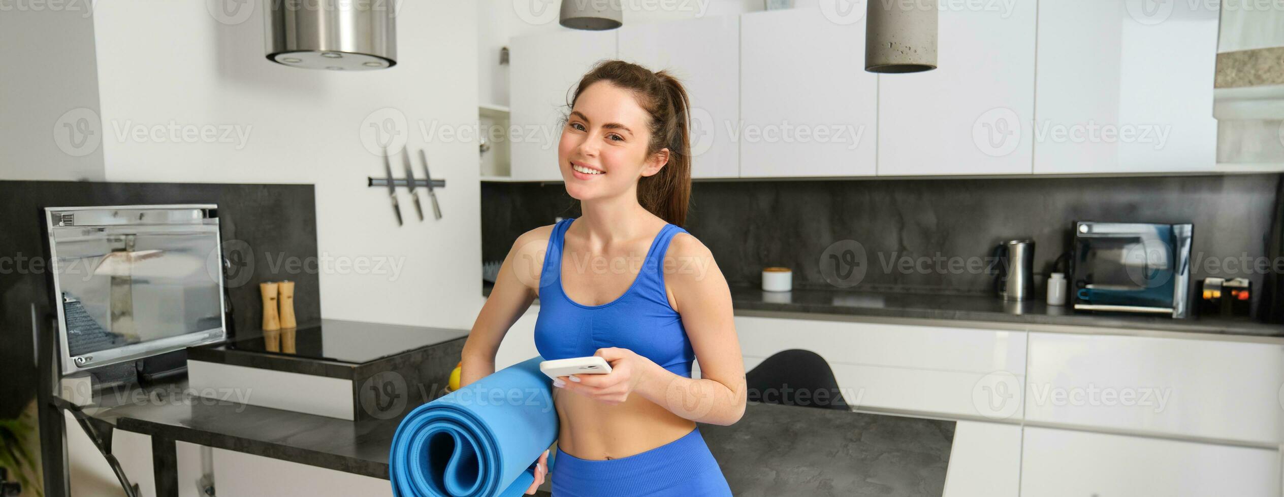 image de fort et en forme aptitude instructeur, femme permanent à Accueil avec caoutchouc tapis et téléphone intelligent dans mains, a un app pour faire des exercices à l'intérieur photo