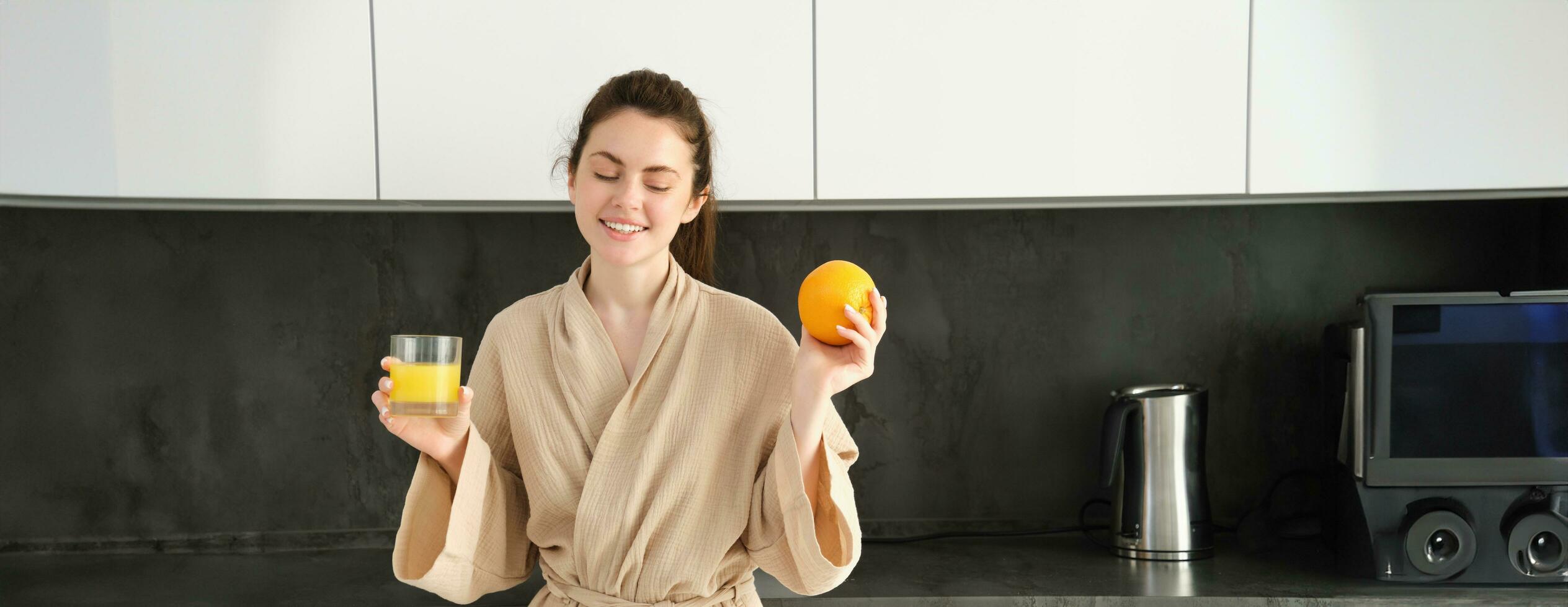 image de beau en bonne santé femme dans peignoir de bain, en buvant Frais jus, montrant Orange fruit, posant dans cuisine photo