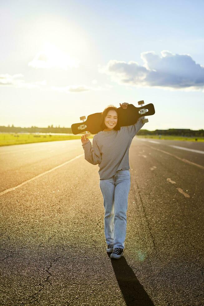 Jeune asiatique femme permanent avec longboard sur ensoleillé route, patinage dans patin parc sur sa croiseur photo