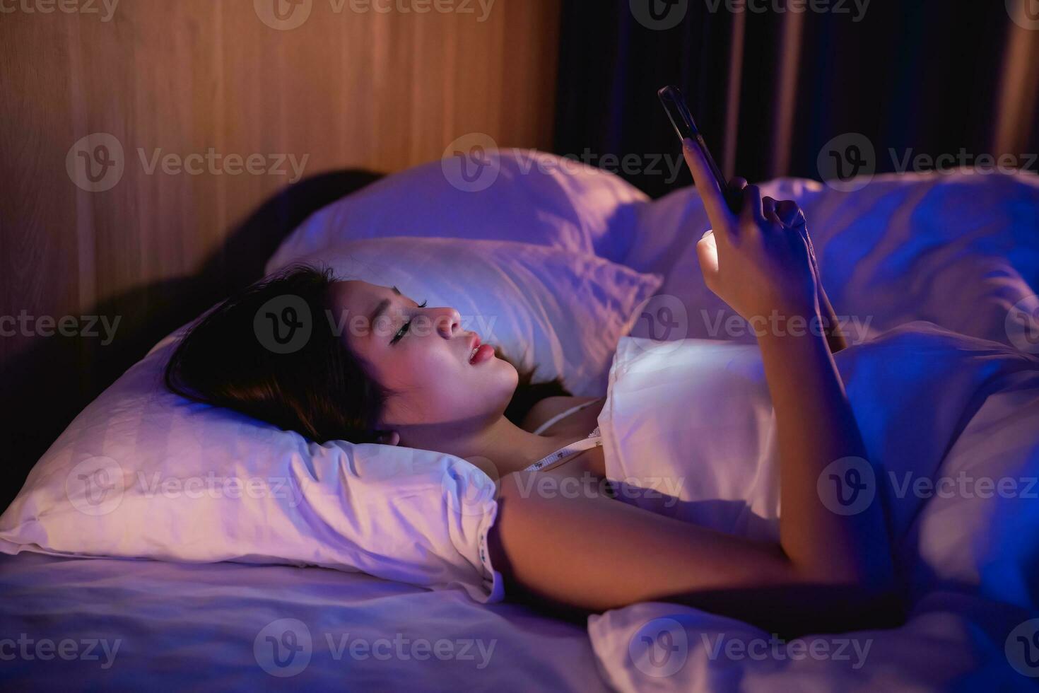 asiatique femme en utilisant mobile téléphone téléphone intelligent pose sur le lit dans le chambre à coucher. somnolent épuisé, pouvez ne pas dormir. insomnie, dépendance concept. femmes défilement social réseaux sur mobile foncé chambre à coucher. photo