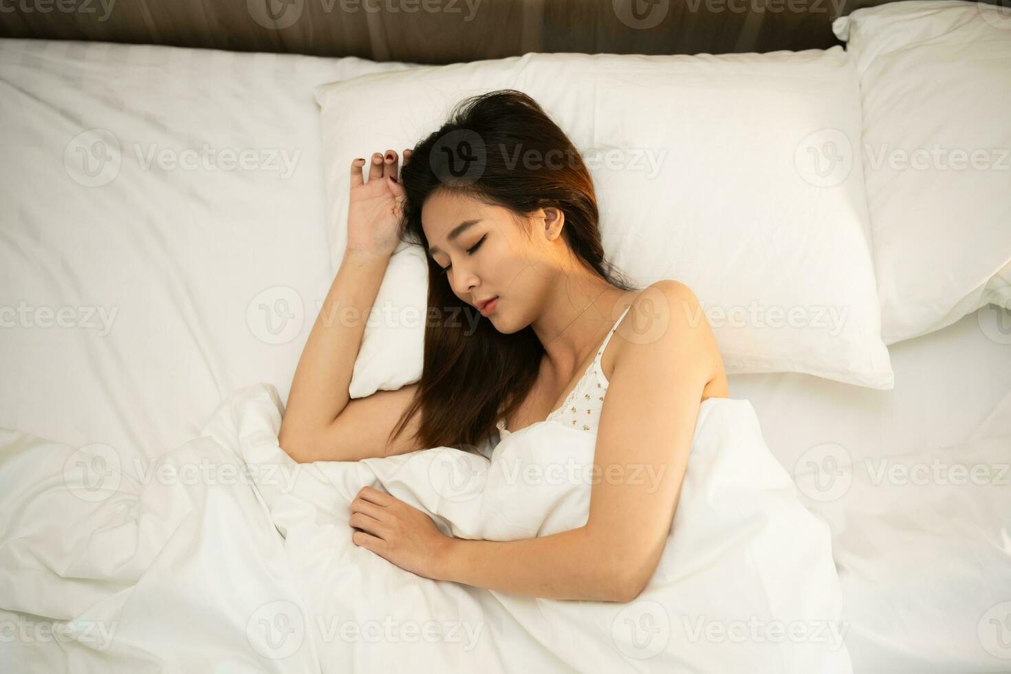 asiatique femme en train de dormir dans confortable lit avec soyeux linge de maison dans le Matin. femmes mensonge dans lit et en gardant yeux fermé tandis que couvert avec couverture. contenu et pacifique, de bonne humeur et confortable. photo