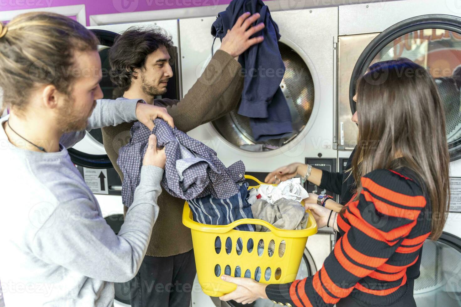 Jeune groupe de millénaire copains est en mettant vêtements dans le la lessive machine photo