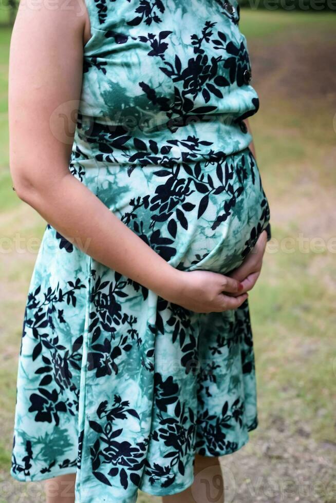 une Enceinte Indien Dame pose pour Extérieur grossesse tirer et mains sur ventre, Indien Enceinte femme met sa main sur sa estomac avec une maternité robe à société parc, Enceinte à l'extérieur maternité tirer photo