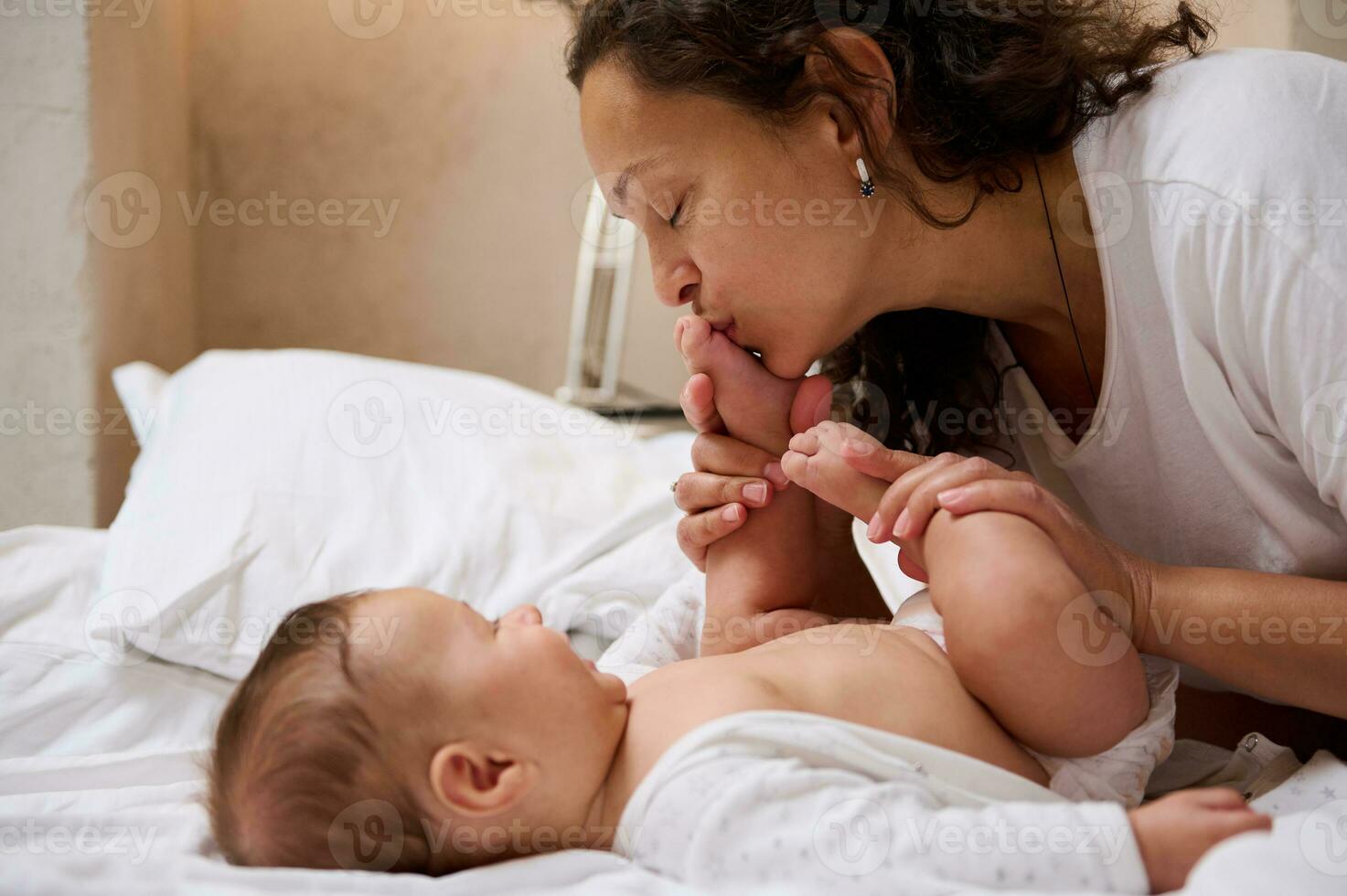 adorable bébé garçon sourit tandis que le sien aimant mère embrasser le sien peu pieds et orteils, profiter content des moments ensemble photo