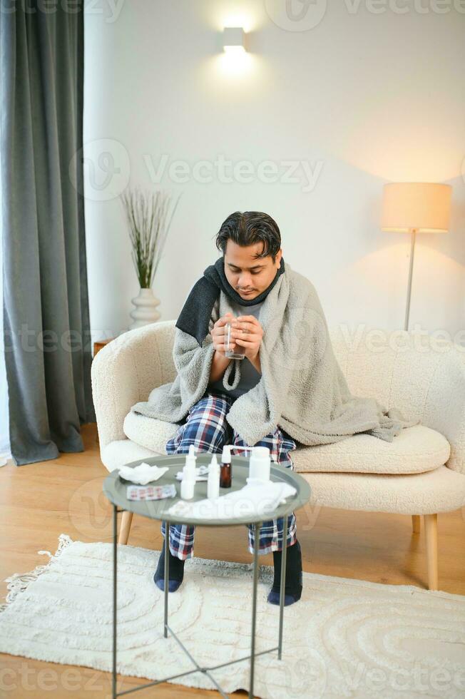 malade homme séance à Accueil sur canapé dans vivant chambre, éternuements a allergie et qui coule nez, Indien homme avec serviette de table a une du froid photo