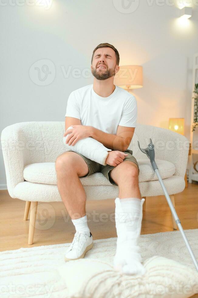 homme récupération de accident fracture cassé OS blessure avec jambe attelles dans jeter cou attelles collier bras attelles fronde soutien bras dans vivant chambre. social Sécurité et santé Assurance concept photo