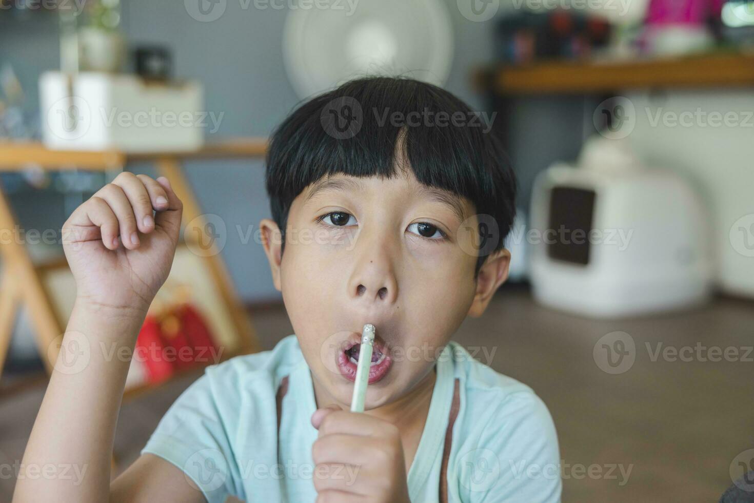 proche en haut portrait de asiatique garçon avec noir frange, noir yeux avec une souriant visage portant une lumière vert chemise Regardez à caméra et séance sur le sol de le sien maison et main en portant crayon à dessiner. photo
