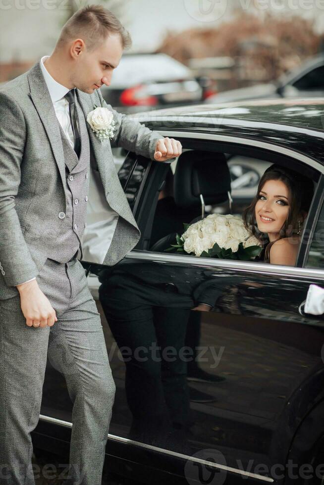 mariage photo. le la mariée avec une bouquet de des roses est assis souriant dans une noir voiture, le jeune marié des stands penché sur le voiture et regards à le la mariée. magnifique maquillage et cheveux. une élégant homme photo