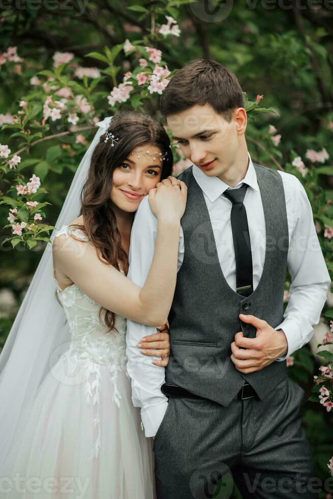 content Jeune couple permanent près magnifique fleurs. le la mariée doucement étreint sa jeune marié derrière le épaules. le Jeune embrasse tendrement, à la recherche bas. mariage portrait. printemps mariage photo