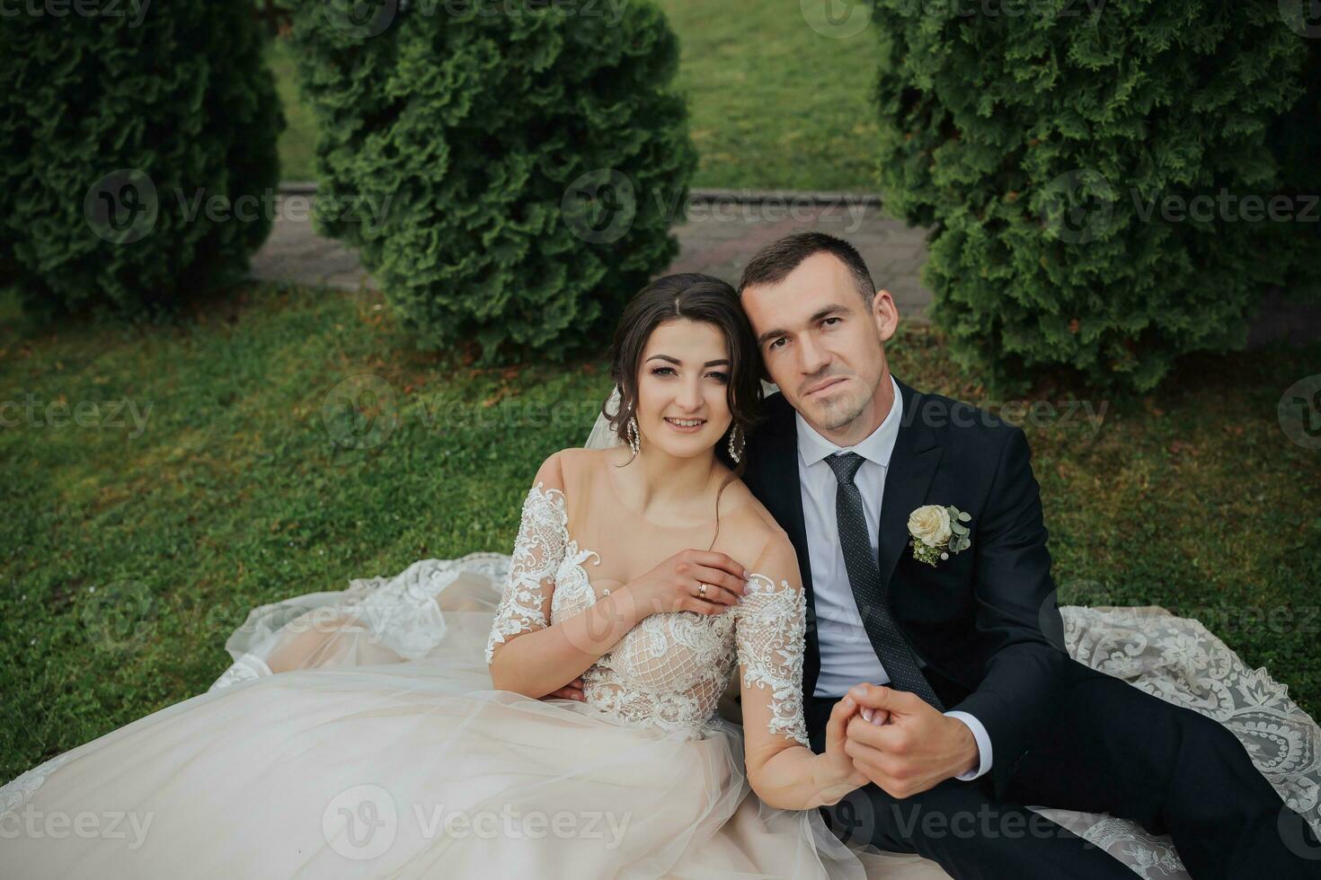 portrait de le la mariée dans la nature. une brunette la mariée dans une blanc dentelle robe et une élégant jeune marié dans une noir costume sont séance sur le herbe, poser, en portant une bouquet. magnifique se maquiller. ouvert épaules. photo