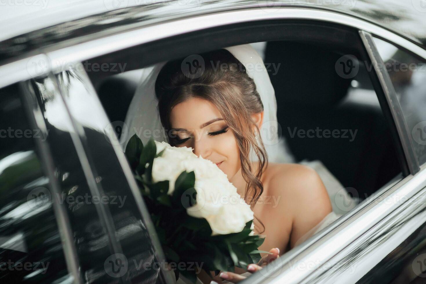 une brunette mariée, souriant sincèrement, est assis dans une noir voiture sur sa mariage journée avec une bouquet de blanc des roses. portrait de le la mariée. magnifique boucles. magnifique maquillage et cheveux. une luxueux blanc robe photo