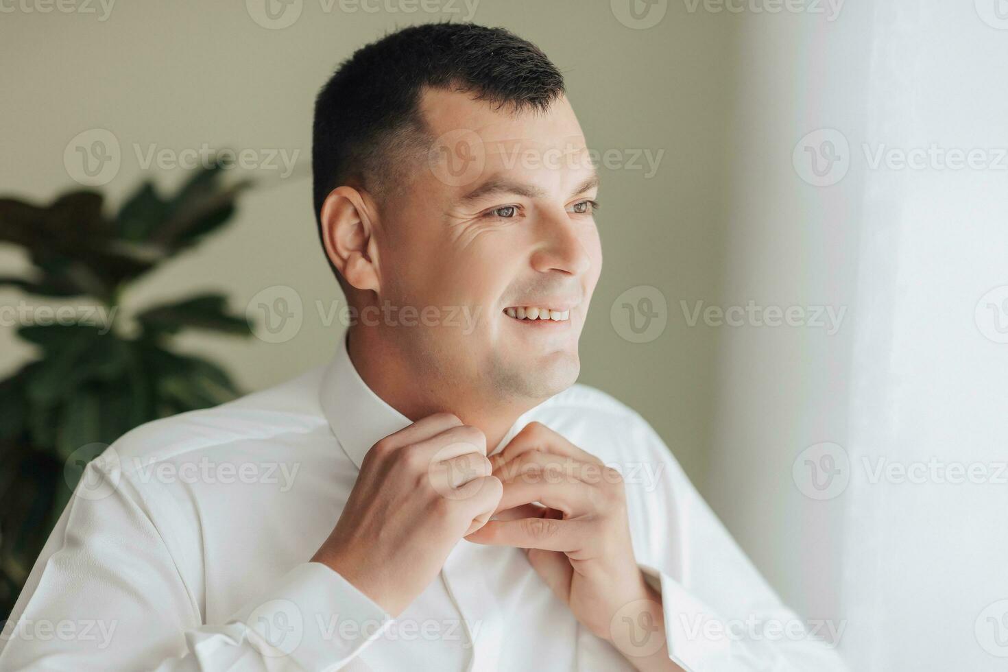 portrait. une Jeune élégant homme dans une blanc chemise pose à la recherche à le côté et attache une bouton sur le sien chemise. mode et style. affaires photo