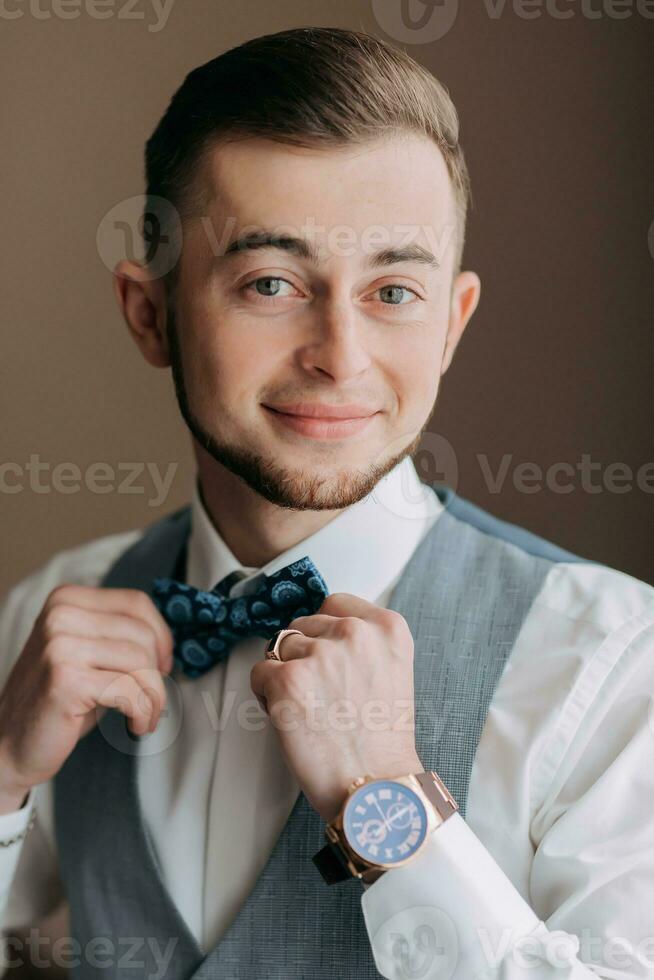 une homme dans une blanc chemise et une gris gilet est posant par le fenêtre et à la recherche à le caméra, ajustement le sien bleu arc cravate. de face voir. une élégant montre. Pour des hommes style. mode. affaires photo