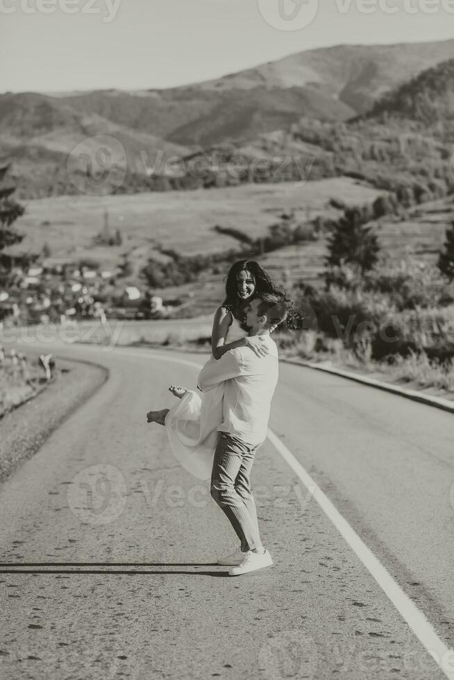 une élégant jeune marié dans une blanc chemise et une mignonne brunette la mariée dans une blanc robe sont encerclant sur un asphalte route contre le Contexte de une forêt et montagnes. noir et blanc photo