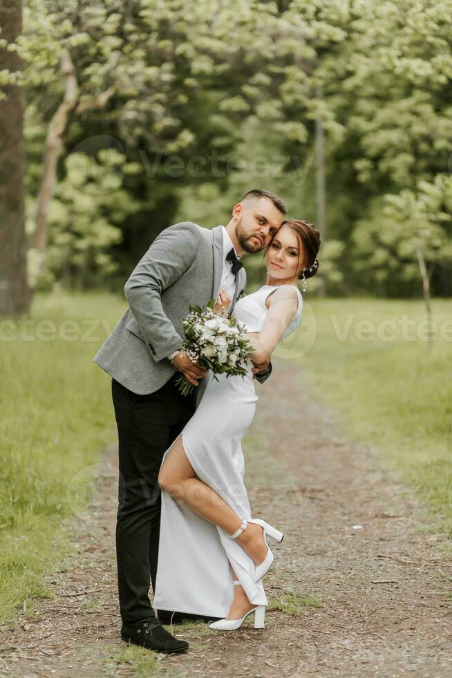 le la mariée dans une blanc soir robe pose avec le jeune marié avec une magnifique coiffure, le jeune marié doucement enveloppements le la mariée et penche contre son. portrait photo